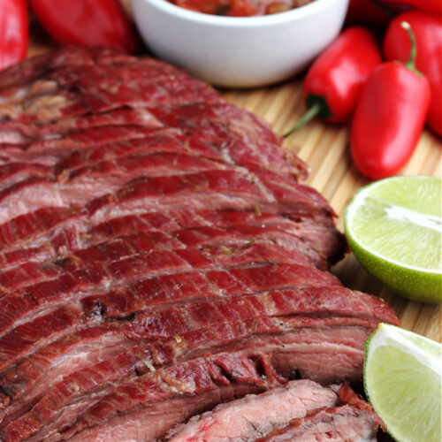 smoked flank steak sliced on a board with limes, peppers and salsa