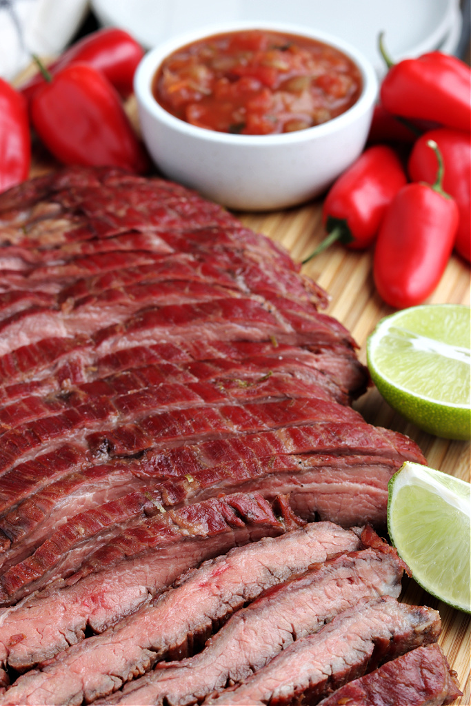 smoked flank steak sliced on a board with limes, peppers and salsa