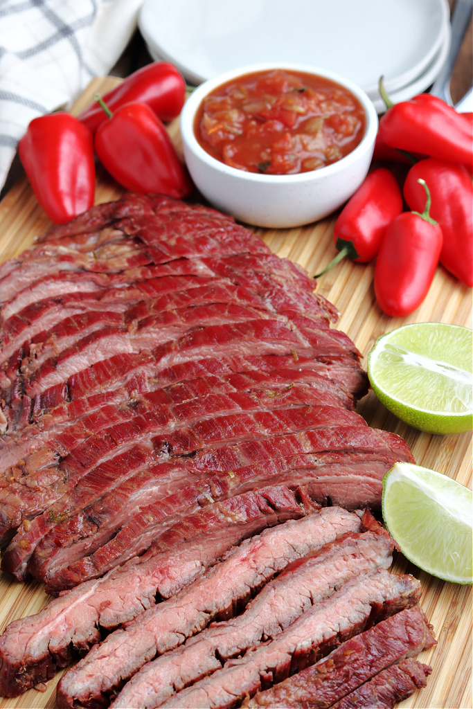 smoked flank steak sliced on a cutting board with limes, red peppers and salsa